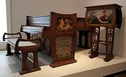 Photo en couleurs d'un piano, un banc et un meuble haut en bois foncé avec un décor peint