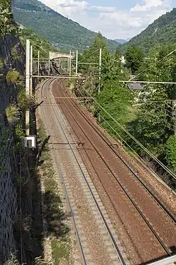 Une ligne de chemin de fer électrifiée à deux voies, avec une portion droite puis une courbe vers la droite, en fond de vallée.