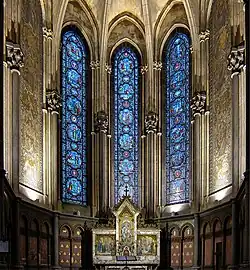 Photographie en couleurs de la chapelle vue de face avec au centre trois vitraux, de part et d'autre une mosaïque murale et en bas le retable.