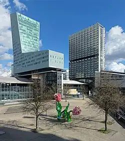 Photographie en couleur d'une vaste place minérale avec en son centre une sculpture représentant des tulipes colorées encadrée de deux arbres sans feuilles et, derrières, deux tours, la première, à gauche, en forme de chaussure de ski, la seconde, plus loin à droite, de forme parallélépipédique