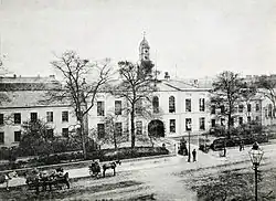 A black and white photograph, showing a stately-looking two-storey building with white walls, extending out of shot to the left and right, with an arched cart entrance at the centre. A modest clocktower rises above the entrance, and the building is surrounded by neat shrubbery and iron railings. A wide street crosses left-right outside of the fence, with a handful of horse-drawn carts and pedestrians in 19th-century clothing.