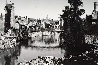 Photographie de maisons détruites à Lisieux le long d'un fleuve avec une passerelle intacte au milieu de la prise de vue