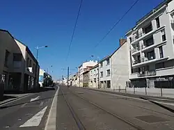 Rue bordée d'immeubles de trois ou quatre étages, des rails de tramway parcourent le centre de la voie