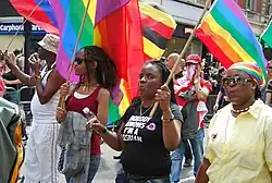 Photographie de plusieurs femmes noires à la Pride tenant des drapeaux arc-en-ciel. Une porte un t-shirt « Nobody knows I'm a lesbian&nbsp;».