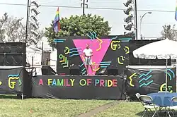 Photographie d'une scène de couleur noir à l'exception d'un grand triangle rose. Il est écrit, en lettres arc-en-ciel "A Family of Pride"