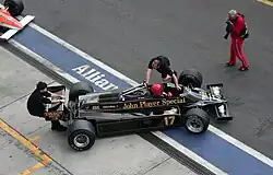 Vue de haut d'une monoplace noire poussée par deux hommes dans la ligne droite des stands.