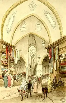 La scène prend place dans un marché couvert. Des passants traversent la rue à pied ou à dos d'âne. De multiples boutiques de tissus ou autres marchandises prennent place de part et d'autres d'un chemin central.