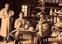 Photographie en noir et blanc de l'intérieur d'un atelier avec trois personnes au travail.
