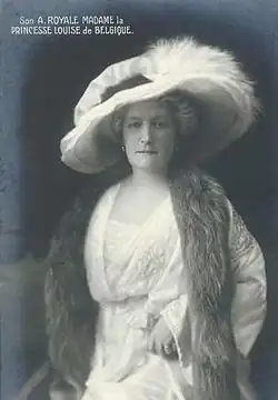 Photographie en noir et blanc d'une femme coiffée d'un grand chapeau à plume, elle porte une robe blanche et une fourrure autour de son cou.
