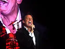 Un homme est tourné vers la droite tout en tenant un micro avec sa main droite.