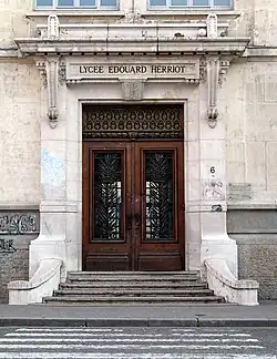Entrée du lycée Édouard-Herriot. Une porte massive en bois, à double battant, au-dessus de laquelle est gravé le nom du lycée, donne sur quelques marches puis le trottoir.