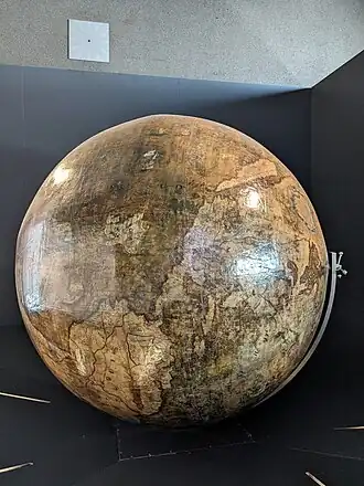 Globe terrestre présenté à l'horizontale sur son axe, centré sur l'Amérique et la Pacifique. Les terres en beige sont parcourues de cours d'eau en noir, les océans sont en vert-gris.