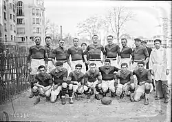 Photographie noir et blanc d'un groupe d'hommes. Au premier plan sept hommes en maillot et short sont à genou ou en position accroupie. Devant le cinquième est posé au sol un ballon ovale. Un homme vêtu d'une veste et d'un pantalon se tient debout à droite de l'image. Derrière ces huit hommes, dix hommes sont debout dont huit sont en tenue de sport. Derrière les personnes à gauche de l'image se trouve un immeuble de plusieurs étages. Quelques arbres poussent derrière au loin.