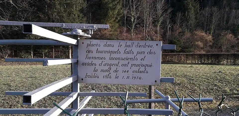 Plaque blanche apposée au tourniquet en métal et indiquant : « Placés dans le hall d’entrée, ces tourniquets faits par des hommes inconscients et avides d’argent ont provoqué la mort de 144&nbsp;enfants brûlés vifs le 01.11.1970 »