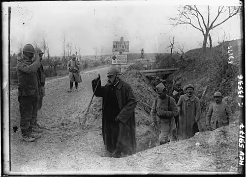 Photo en noir et blanc d'une personne aux cheveux blancs, à la moustache foncée et avec un casque d'artilleur sortant d'une tranchée et saluant deux militaires