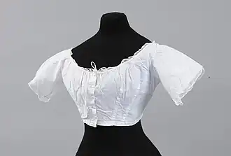 Cache corset pour crinoline en cotonnade blanche. Décolleté forme bateau, petits mancherons soulignés de dentelle à l’aiguille. Boutonnage milieu devant par 5 boutons recouverts et une agrafe.   Vers 1860.