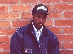 portrait  de l'artiste afro-américain avec veste bleu et casquette noir devant un mur de brique rouge