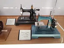 Deux machines à coudre de taille réduites sont exposées dans une vitrine. Celle de devant est peinte en bleu-gris, celle de derrière est noire. Toutes les deux sont conçues pour être actionnées à la main.