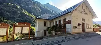 Photographie de la mairie d'Orelle en été avec des fleurs au centre dudit chef-lieu, il y a des panneaux municipaux avec l'inscription d'entrée révélant que le bâtiment est la mairie.