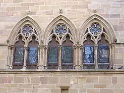 Photo couleur d'un groupe de trois fenêtres géminées en ogive double à colonnettes. Les baies sont munies de panneaux de bois dotés de vitraux en losange.
