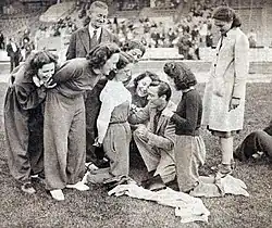 Agenouillé au sol, entouré de femmes, un homme écrit sur les vêtements de l'une d'entre elles avec un stylo.