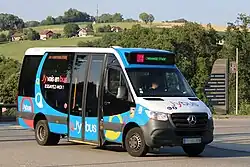 Photographie en couleurs d’un minibus bleu, blanc et noir circulant sur la route des Creuses devant un arrière-plan rural.