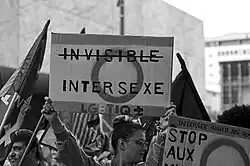 Photographie en noir et blanc d'une personne dans une manifestation brandissant une pancarte en carton avec écrit "invisible" barré suivi de "Intersexe" et "LGBTIQ+", avec le drapeau intersexe en fond