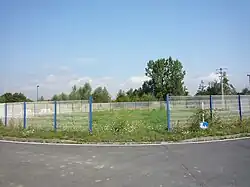 Photographie montrant un rond-point partiellement entouré de grilles blanches et envahi de mauvaises herbes, au centre duquel se trouve la tête de puits matérialisée de la seconde fosse de Marchiennes, ouverte par la Compagnie des Canonniers de Lille.
