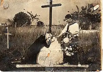 Photo en noir et blanc montrant un homme agenouillé sur une tombe ornée d'une croix, avec un autre homme debout à côté, tenant une couronne de fleurs