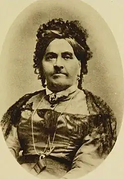 Photographie en noir et blanc d'une femme, les cheveux surmontés d'une coiffe, portant une large robe.