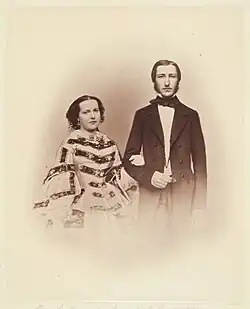 À gauche Marie-Henriette, les cheveux foncés divisés par une raie et portant une robe à crinolines et à passants horizontaux, marquant la taille, tient le bras droit de Léopold, revêtu d'une longue redingote sombre, portant une cravate sombre enserrant un col clair, son visage est recouvert de longs favoris foncés