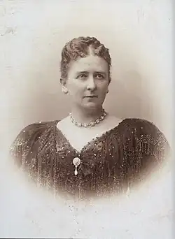 Photographie en buste de Marie les cheveux coiffés vers l'arrière, portant un rang de pierres précieuses entourant son cou et vêtue d'une robe décolletée sombre ornée d'un pendentif central