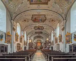 Dans une nef blanche des tableaux au cadre d'or, un plafond crème richement peint de tableaux, une allée centrale bordée de bancs bien rangés.