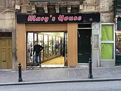 Photo d'un magasin à la devanture beige avec une inscription rouge sur fond noir en haut de celle-ci. Un homme se tient près de l'entrée, composée d'une série de vitrines et d'un renfoncement vers l'intérieur du magasin.