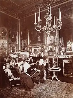 Photographie en noir et blanc d'une Victoria âgée assise à côté d'une jeune femme (Beatrice) lisant un journal. La pièce est richement décorée avec de nombreuses photographies et peintures et un large chandelier est suspendu au plafond.