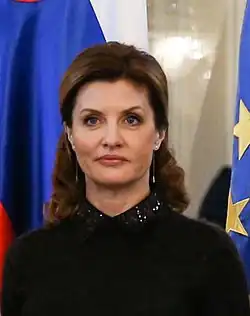Gros plan sur une femme blanche vêtue de noir se tenant face-caméra. Les cheveux de celle-ci sont bruns et ramenés vers l’arrière. En arrière plan, les drapeaux de l’Union européenne et de la Russie sont visibles.