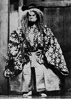 Acteur debout, figé, en tenue traditionnelle avec un masque massif et sévère et une large perruque.