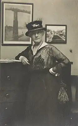 La photo en noir et blanc montre Mattie Edwards Hewitt, portrait de trois quarts de longueur, debout, face à l'avant, bras droit sur la commode, avec un chapeau.