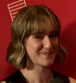 Portrait d'une jeune femme aux cheveux blonds et vêtue en noir. Elle sourit vers l'objectif alors qu'elle est interviewée. Le fond est rouge et l'éclairage tamisé.