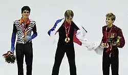 Photographie de trois patineurs en costume sur un podium, une médaille autour du cou, les deux à l'extérieur portant un bouquet de fleurs, l'un au centre entouré du drapeau russe.