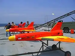 Trois drones rouges fuselés avec rampe sur pont de porte-avion. Les ailes delta et les nez sont jaunes (3 × 2,5 m env.).