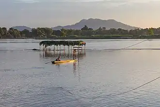 Pirogue du Mékong au coucher du soleil avec deux hommes travaillant sur une ligne électrique pour connecter un abri dans le Si Phan Don) près de Don Puay. Leur but est de créer un stand pour vendre des boissons ici dans quelques jours lorsque le niveau de la rivière aura baissé.