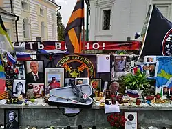 Photo couleur d'un muret recouvert de portraits, de fleurs et de drapeaux.