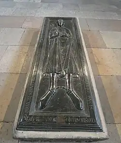 plaque rectangulaire de couleur sombre sculptée figurant un homme couché sur le dos avec une inscription tout autour, sur un socle rectangulaire légèrement surélevé