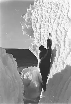 Photographie d'un homme debout, vu de dos, accroché à son piolet sur une paroi verticale.