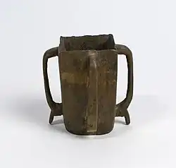 Photo d'un récipient en bois foncé, de section quadrangulaire, à quatre poignées se prolongeant par des pieds à la base.