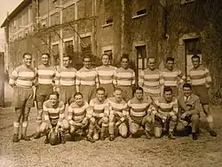 Photo sépia représentant 15 personnes devant un bâtiment en briques des années 1930 avec deux grandes portes.
