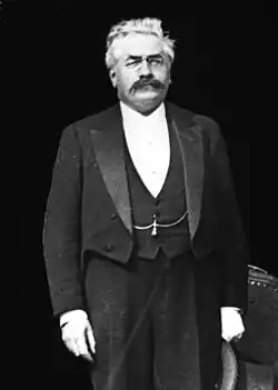 Photo en noir et blanc d'un homme debout, avec les cheveux blancs, une moustache foncée, des lorgnons, un costume trois pièces, un chapeau à la main