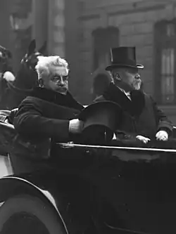 Photo en noir et blanc de deux hommes assis à l’arrière d'une calèche : celui à gauche a des cheveux gris-blancs hirsutes, une moustache foncée, des lorgnons et tient dans sa main droite un chapeau, tandis que celui de droite a une moustache et un bouc blancs, et porte un chapeau sur la tête ; à l’arrière-plan, un garde républicain est distinguable