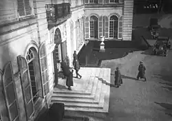 Photo en noir et blanc du perron d’un bâtiment, dans lequel pénètre un individu en civil et un militaire